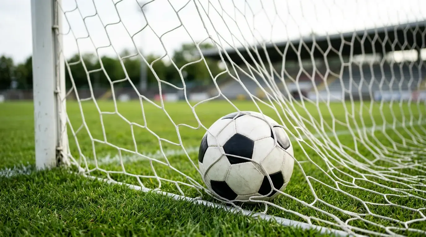 Bola de futebol junto a rede da baliza num relvado verde com luz natural